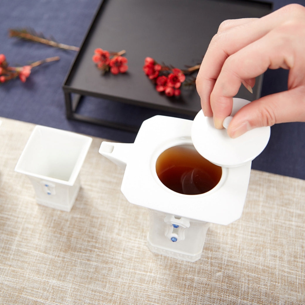 明珠在握白瓷茶壺沖泡情境,熱水注入壺中並搭配方形茶杯與紅色茶席花果