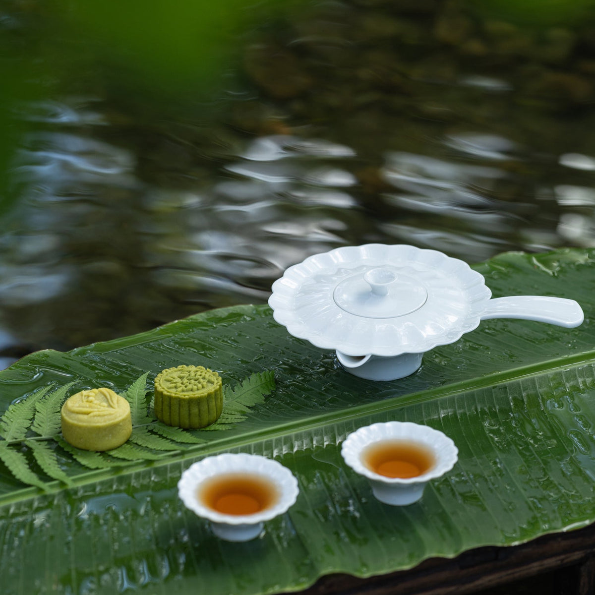 〈山水情深〉茶壺與兩杯熱茶擺在黑色圓盤上，旁有蕨葉與茶點